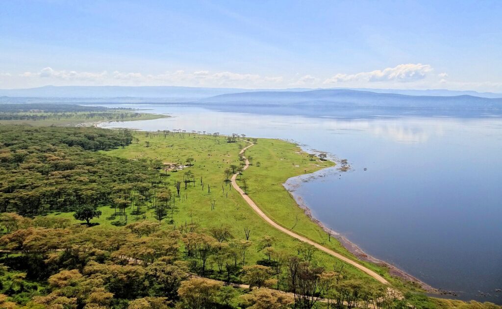 Vista de cima, de Baboon Cliff, sobre a margem do lago, uma trilha que acompanha um campo verde salpicado de animais, bosques de acácias e o lago até as colinas