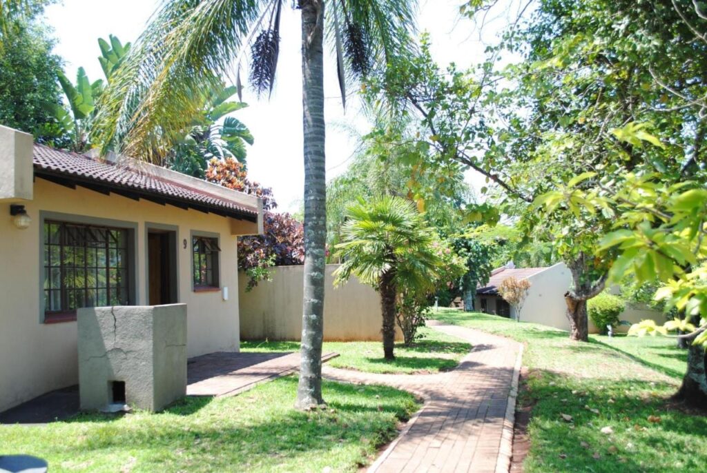 Um chalé dos Eagles Nest Chalets, de paredes rebocadas e telhado de telhas, com sua churrasqueira de concreto, à beira de um caminho de tijolos sob as palmeiras