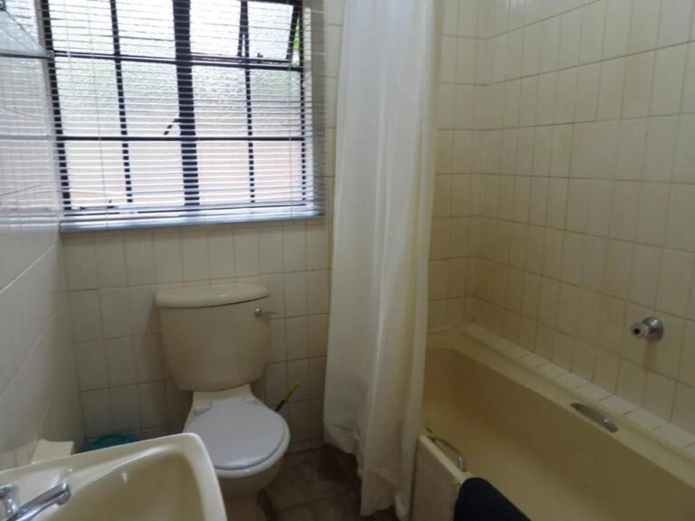 Um banheiro azulejado, com banheira com cortina, vaso sanitário e pia, sob uma janela com persiana