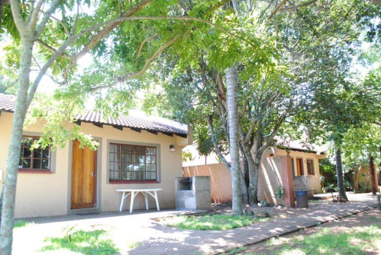 Um chalé sob as árvores, com uma mesa de jardim branca e uma churrasqueira de concreto ao lado da porta de madeira