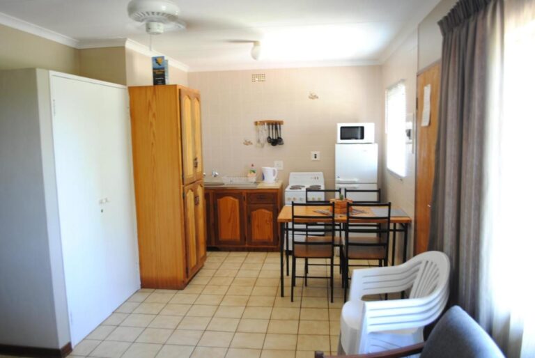 A sala de um chalé, com cozinha compacta, mesa de jantar, guarda-roupa de madeira e uma cadeira branca