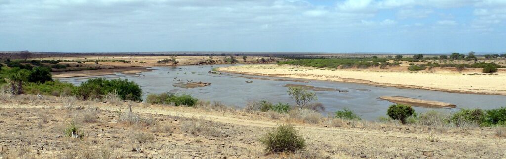O rio Galana, largo e marrom, faz uma curva entre bancos de areia clara e arbustos, sob um céu nublado