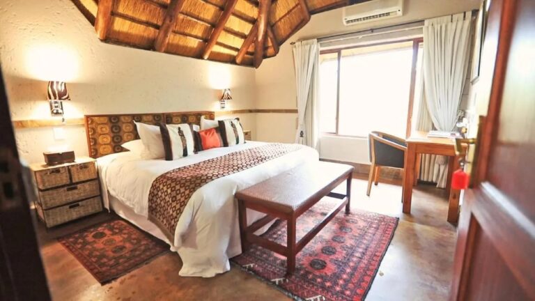 Um quarto sob uma estrutura de palha, com cama de casal, banco de madeira e tapetes vermelhos