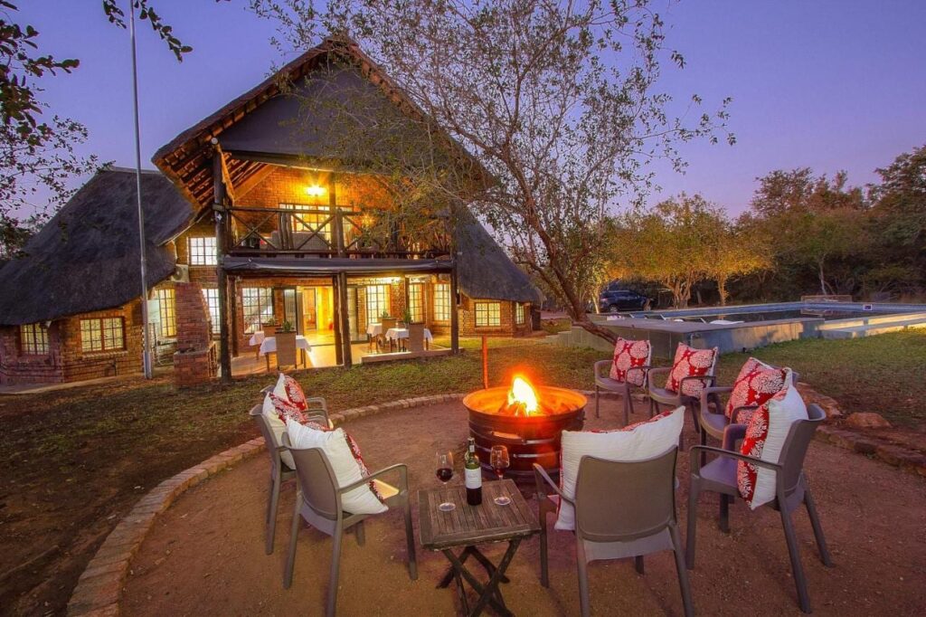 O prédio de tijolo e palha do Imbube Safari Lodge ao anoitecer, com um braseiro aceso cercado de poltronas, perto da piscina