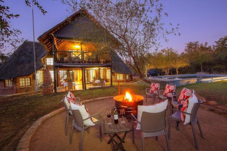 O prédio de tijolo e palha do Imbube Safari Lodge ao anoitecer, com um braseiro aceso cercado de poltronas, perto da piscina