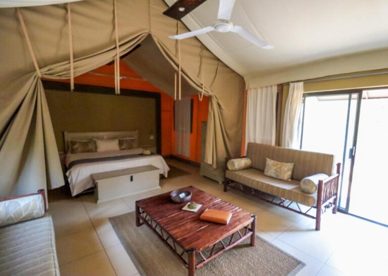 O interior de uma tenda, com cama de casal, sofá, mesa de centro de madeira e uma grande porta de vidro