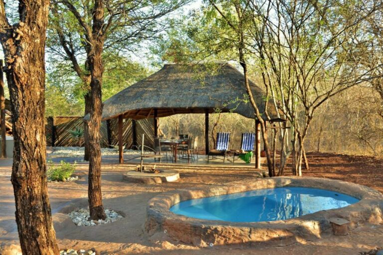 Uma piscina redonda diante de um abrigo com telhado de palha, com mesa de jantar e duas espreguiçadeiras listradas
