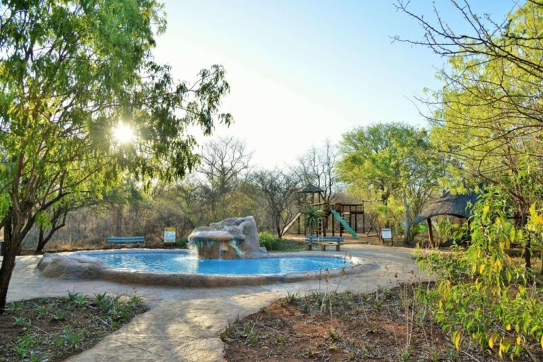 Uma piscina com cascata de pedras, diante do playground e de um abrigo de palha, contra a luz