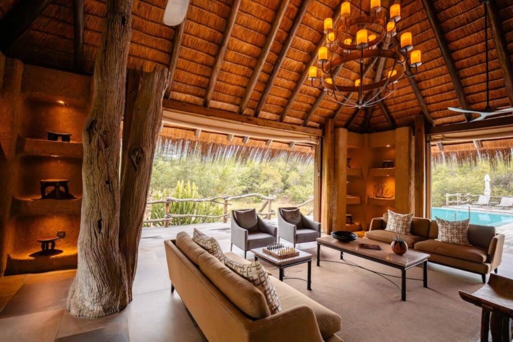 A sala de estar do Kapama River Lodge sob um alto telhado de palha, aberta para uma piscina e a mata