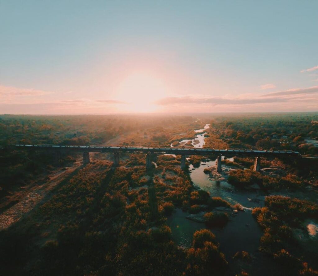 O trem do Kruger Shalati sobre a ponte Selati, visto do alto, acima do rio e da mata, com o sol baixo no horizonte