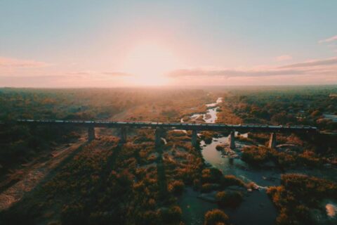O trem do Kruger Shalati sobre a ponte Selati, visto do alto, acima do rio e da mata, com o sol baixo no horizonte