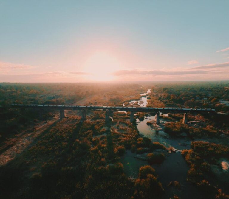 O trem do Kruger Shalati sobre a ponte Selati, visto do alto, acima do rio e da mata, com o sol baixo no horizonte
