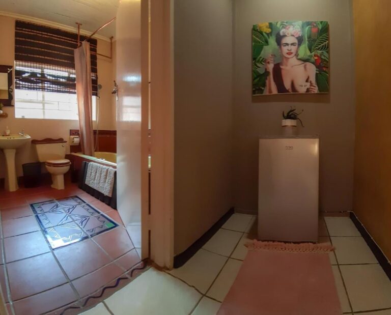 Um banheiro com banheira, pia e vaso sanitário, visto de um corredor onde uma pequena geladeira fica sob um retrato de mulher