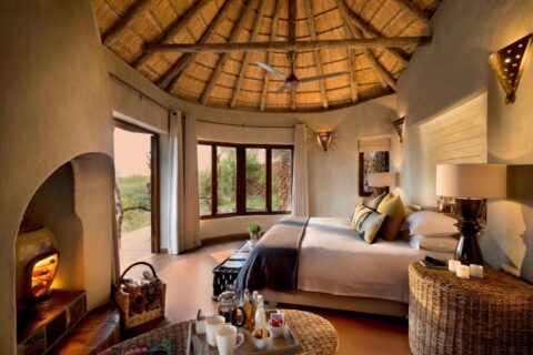 Uma suíte do Madikwe Safari Lodge sob um telhado de palha, com cama de casal e a lareira acesa