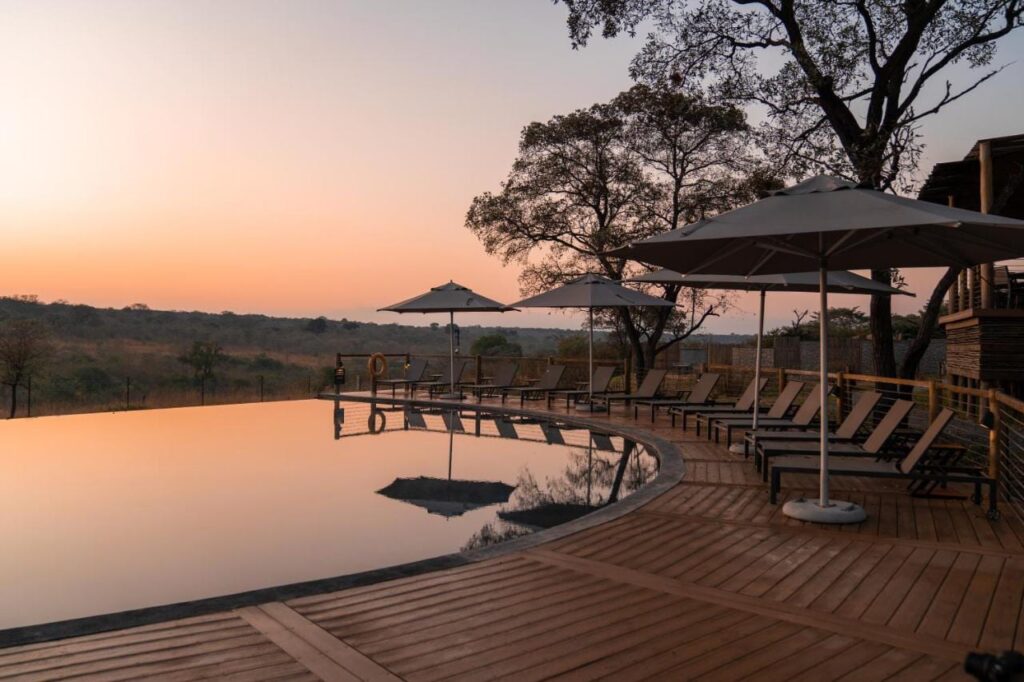 A piscina de borda infinita do Mdluli Safari Lodge sob um céu alaranjado, cercada de espreguiçadeiras e guarda-sóis