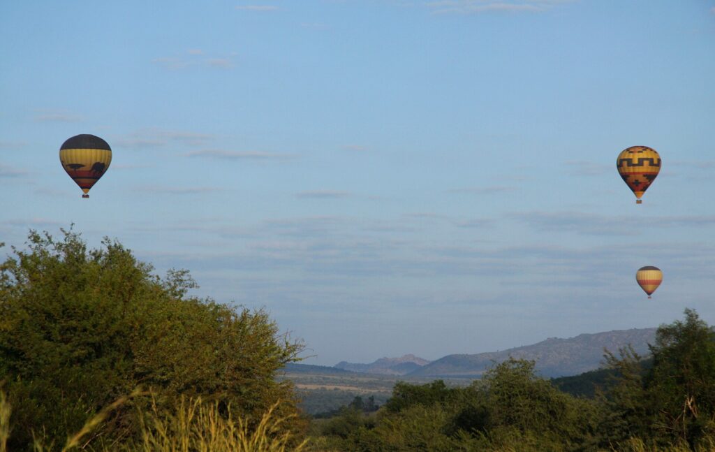 Três balões amarelos e pretos flutuam em um céu azul-claro sobre a savana verde, com as colinas da cratera no horizonte