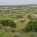 Vista de Observation Hill sobre uma planície verde manchada de sal, uma trilha vermelha, três 4x4 estacionados perto de um pequeno prédio ocre e colinas baixas ao longe