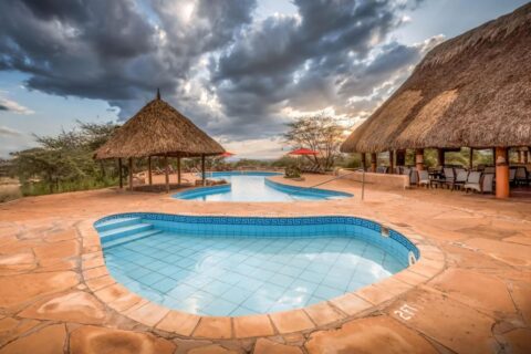 Uma pequena piscina de azulejos em primeiro plano e a piscina grande do Samburu Sopa Lodge, entre um quiosque de palha e um terraço com telhado de palha