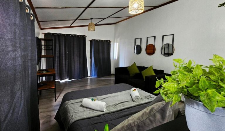 Uma cama de casal cinza com toalhas enroladas, um sofá preto e cortinas escuras, em um estúdio com teto quadriculado de madeira