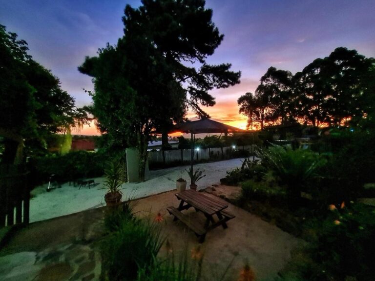 O jardim ao pôr do sol, com uma mesa de piquenique sob árvores grandes e um céu alaranjado
