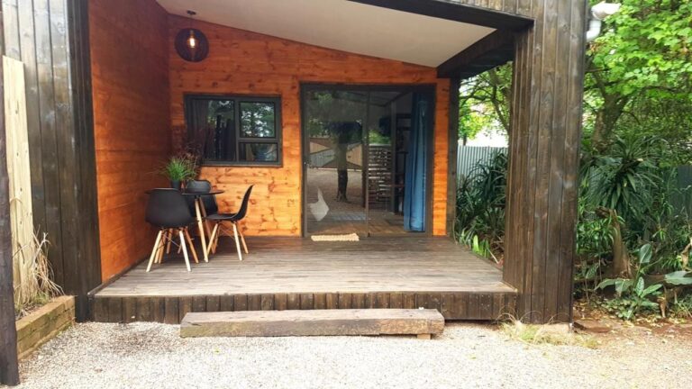 O deck de madeira de um chalé, com uma mesa e duas cadeiras pretas diante de uma porta de vidro de correr