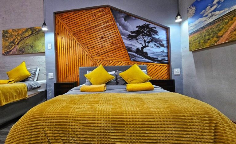 Uma cama de casal com manta amarela diante de uma cabeceira de ripas de madeira e de uma pintura de árvore em preto e branco, ao lado de uma segunda cama