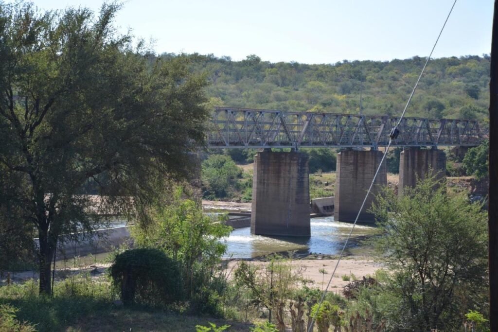 Uma ponte metálica sobre pilares de concreto acima do rio Olifants, vista do jardim do Three Bridges B&B