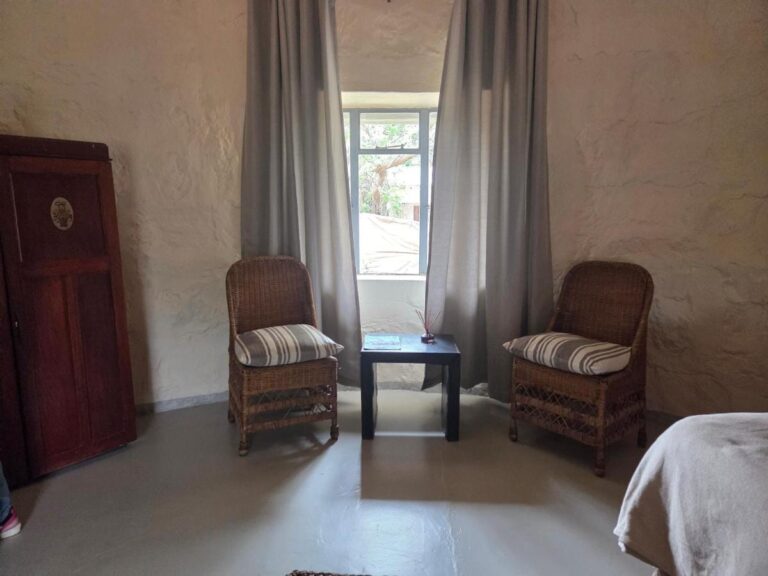 Duas poltronas de rattan e uma mesinha diante de uma janela com cortinas cinza, ao lado de um guarda-roupa de madeira