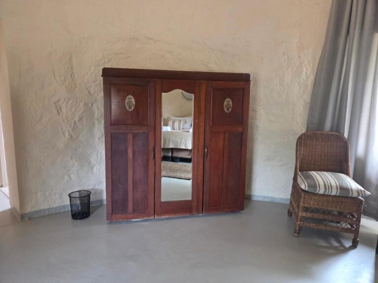 Um guarda-roupa de madeira escura com porta espelhada e uma poltrona de rattan, em um quarto de paredes rebocadas