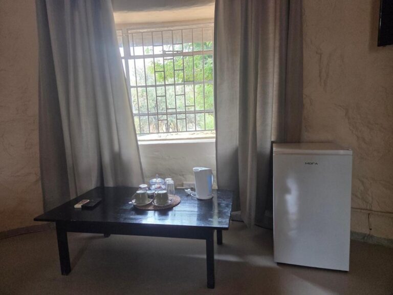 Uma mesa de centro com xícaras e chaleira diante de uma janela telada, ao lado de uma pequena geladeira